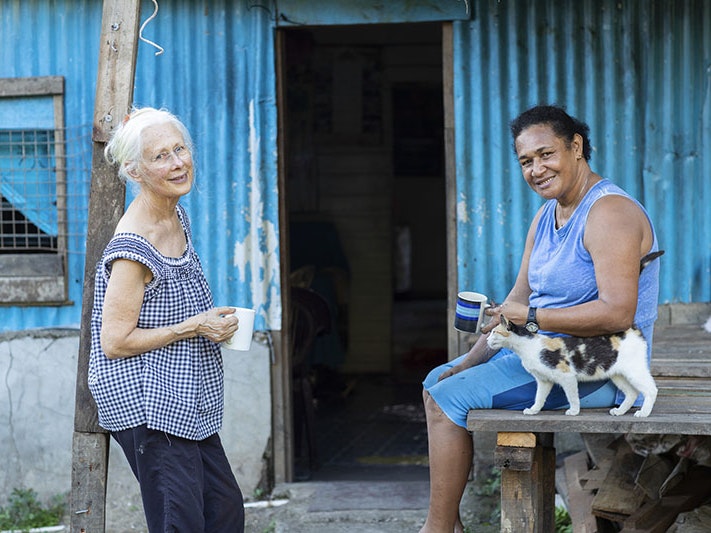 Robin White (left) and Tamari Cabeikanacea (right) in Fiji, 2019. Photo by Michael O’Neill. Te Papa Two women standing in front of a blue corrugated iron shed. They are both holding cups of tea and looking at the camera. There is a cat beside one of them.
