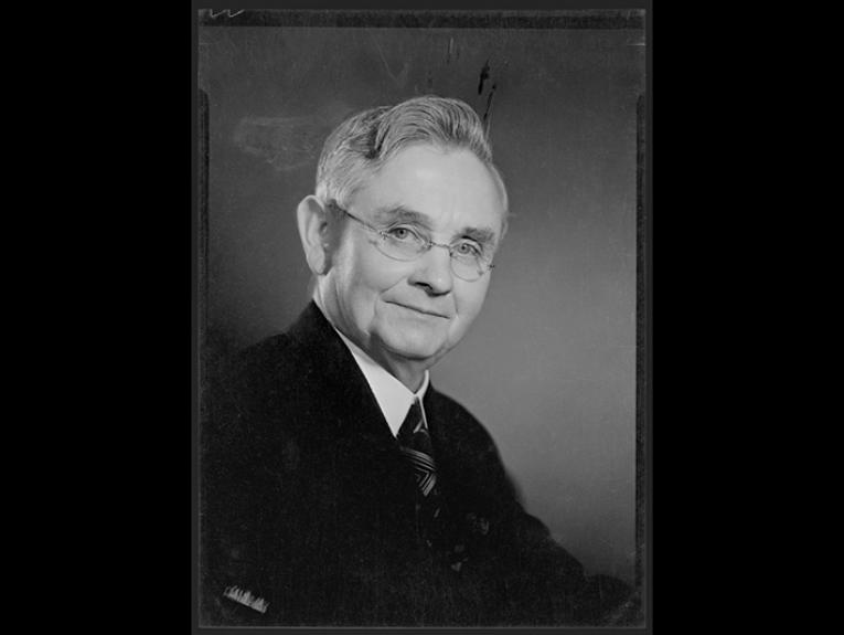 A head and shoulders portrait of a man in a dark suit and glasses.