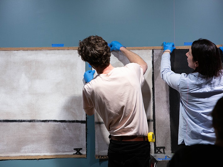 Te Papa staff installing Walk (Series C), 1973 by Colin McCahon, 2019. Photo by Jack Fisher. Te Papa (144200)    Two people with their backs to the camera and wearing blue gloves are hanging a large painting on the wall.