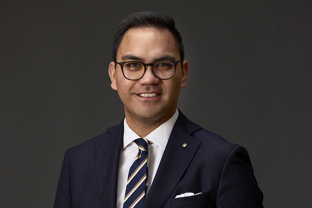 Head and shoulders of a man in a jacket and tie against a dark background. He is wearing glasses.