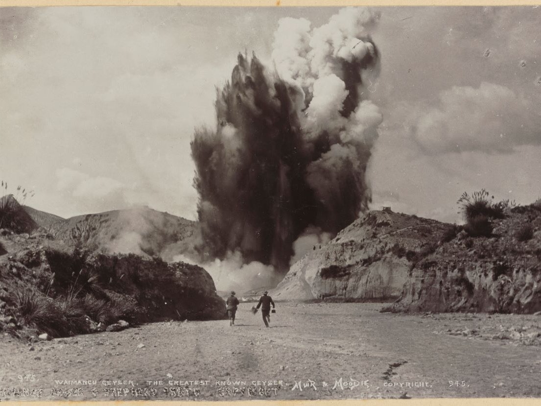 Waimangu Geyser, the greatest known geyser A massive geyser erupting against a hilly horizon. Two small figures are seen in the foreground.