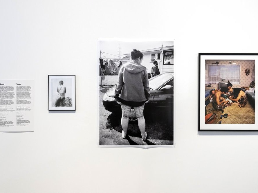 Installation view, Then and There, Here and Now: Samoan Portraits, 14 Sept–10 Dec 2023, New Zealand Portrait Gallery Te Pūkenga Whakaata. Photo courtesy of the New Zealand Portrait Gallery A photo of an exhibition wall with three photos on it of people either with traditional tattoos or receiving one.