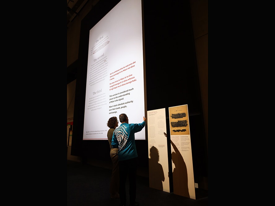Signs of a Nation, 2023. Photo by Tia Nepia Su'a. Te Papa A dark room with a large white projection with text on it. There are two people standing in front of the projection looking at the text.