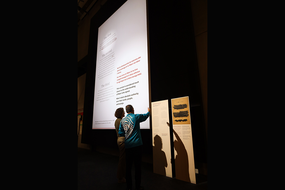 A dark room with a large white projection with text on it. There are two people standing in front of the projection looking at the text.