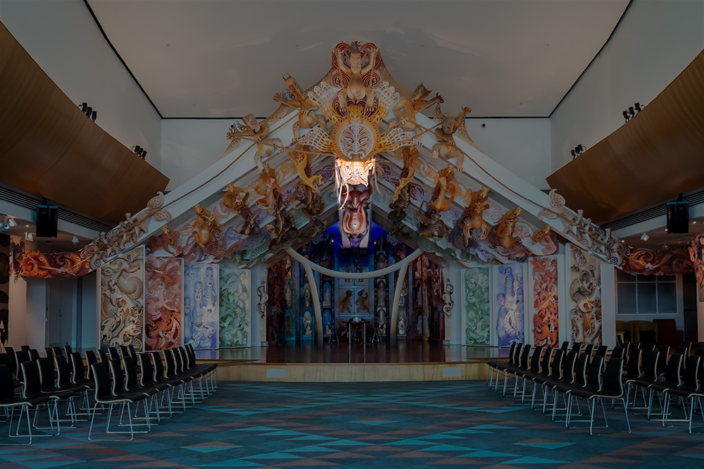 A multicoloured stage with an open-wall house at the back with carvings made of MDF on the sides and rafters. There are chairs either side of the stage set out so they face each other.  One section is highlighted the rest of the image is darkened.