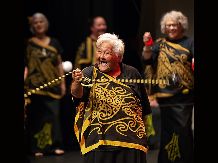 Matariki Festival 2018. Ngāti Whātua Ōrākei Te Puru O Tāmaki Taikura performing at Taikura Kapa Haka event, 2018. Photograph by Jo Moore. Te Papa (108674) An older woman in a yellow and black top is spinning long poi on stage with other older people.