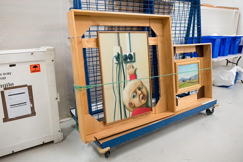 A rack on wheels in a store room – the rack has two art paintings tied on with strops.