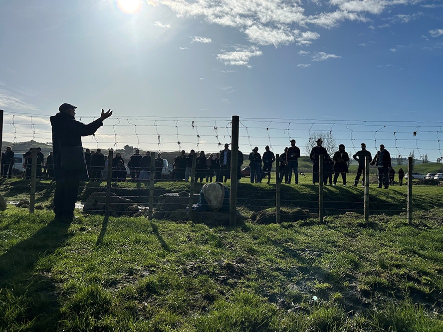 Whaikōrero during the reburial ceremony, July 2023. Image: Amber Aranui 2023. People standing on a grassy field with a fence. One person is talking and gesturing to the people. It is early morning and the shadows are long.