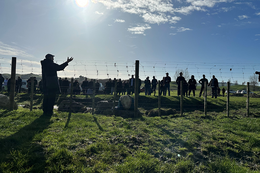 People standing on a grassy field with a fence. One person is talking and gesturing to the people. It is early morning and the shadows are long.