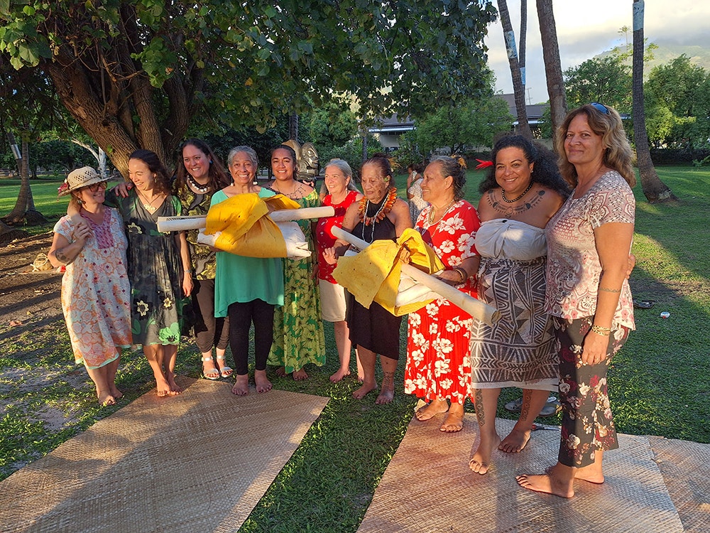 Tapa makers with the completed bundles to go to resides with Te Papa and the other with Te Fare Iamanaha-Musée de Tahiti et des Îles. Photo by Rebecca Rice. Te Papa Several women standing in a group in the late afternoon sun. Some of them are holding bundles of tapa.