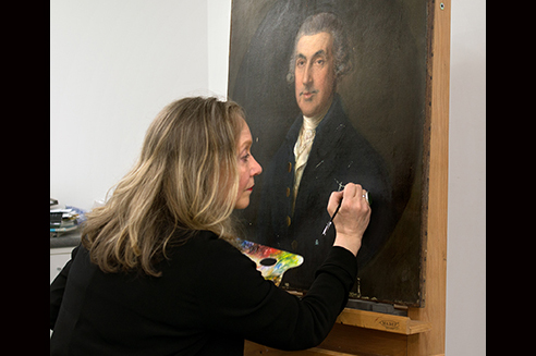 A woman is touching up an old painting on an easel in a studio.
