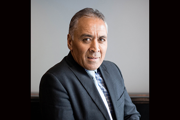 A photo of a man in a suit leaning one arm on a table and looking at the camera