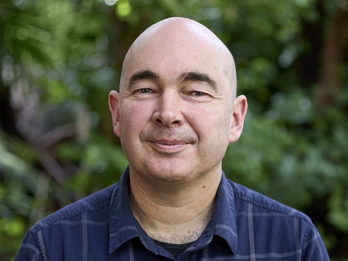 Leon Perrie. Photo by Yoan Jolly. Te Papa Head and shoulders photo of a man with no hair smiling at the camera.