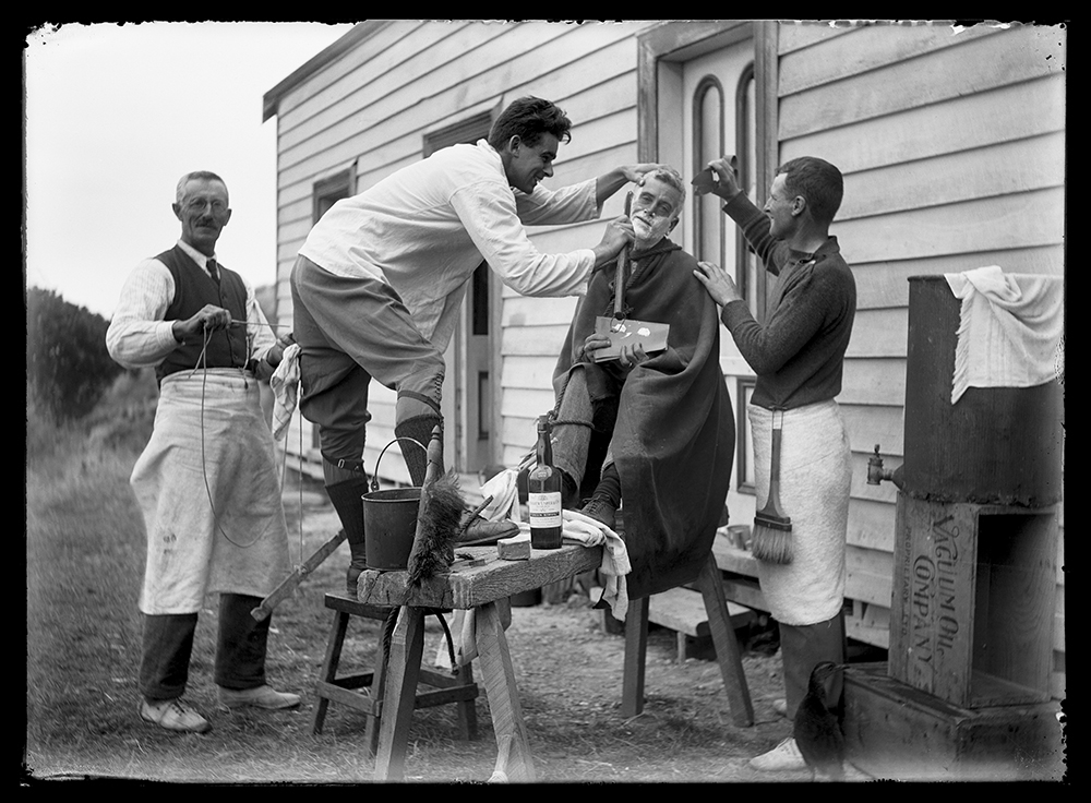 A black and white photo of four men outside a house. One of them is sitting in a tall chair and being shaved by another man.