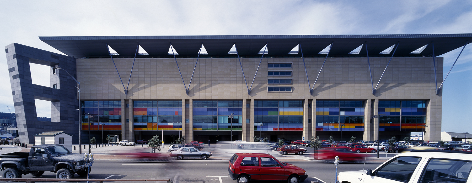 A photograph of the side of a building that has a lot of different coloured glass panels on the lower half of the wall.  There are cars passing in the foreground.