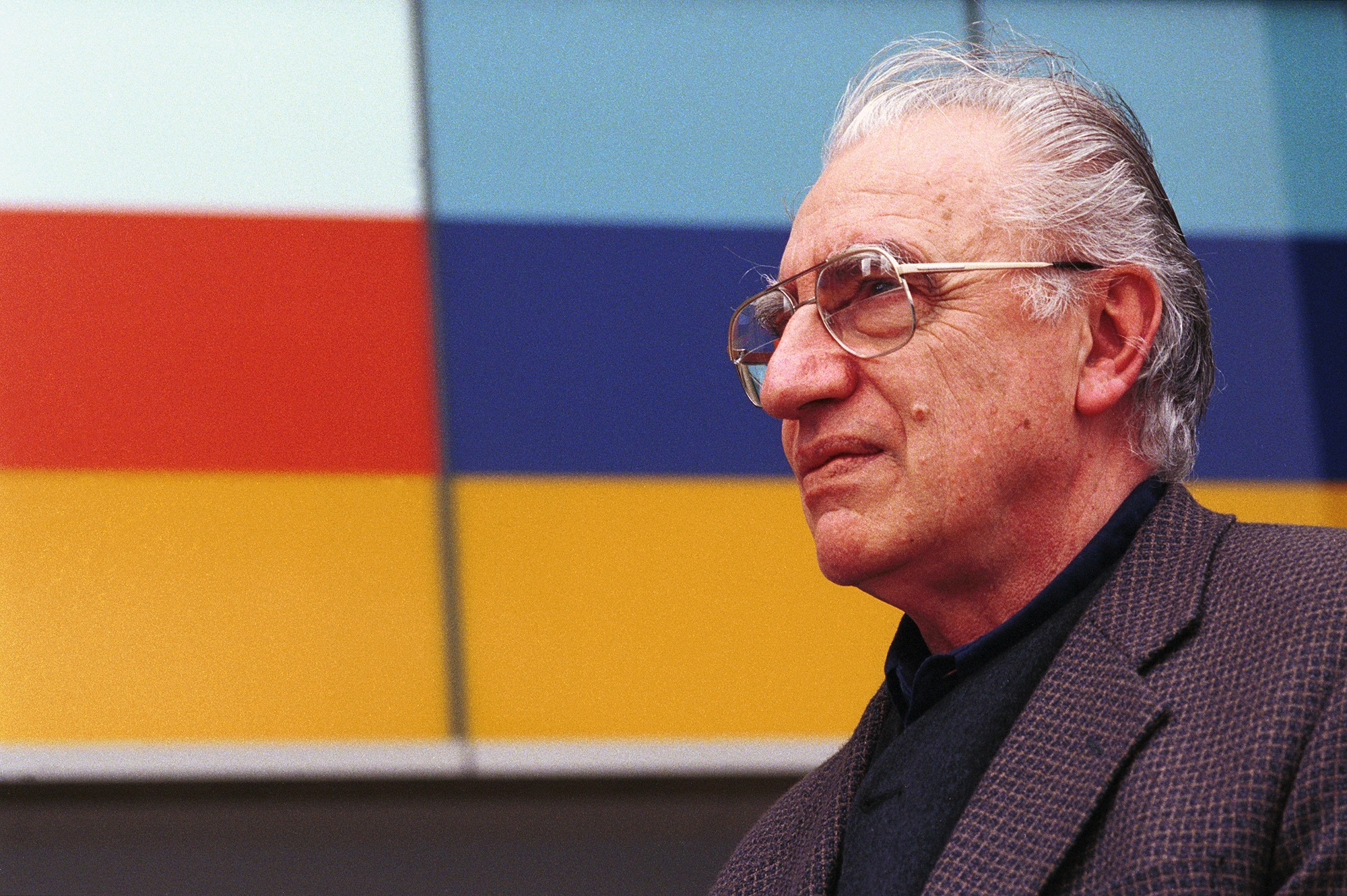 A side profile of a man's head and shoulders. He is standing infront of a colourful building