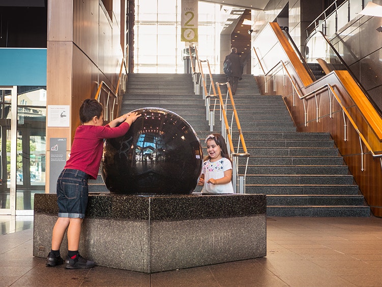 Photo by Johnny Hendrikus. Te Papa (136706) Two young children have their hands on a big black ball made of stone set in a stone box. The ball has water on it.