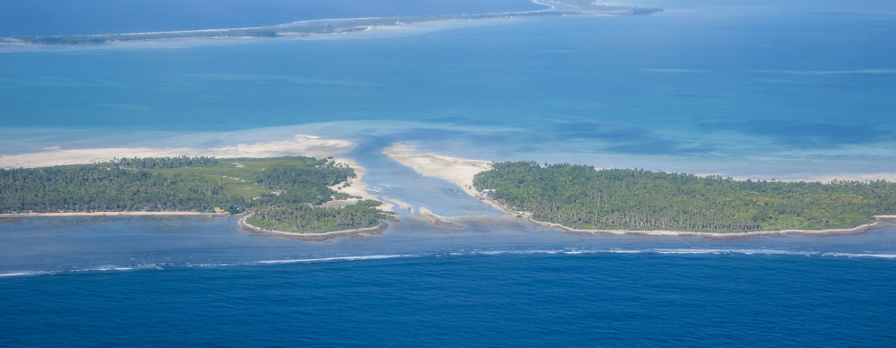 An aerial photo of two parts of an island in the Pacific ocean. There is a small strip of an island in the distance.