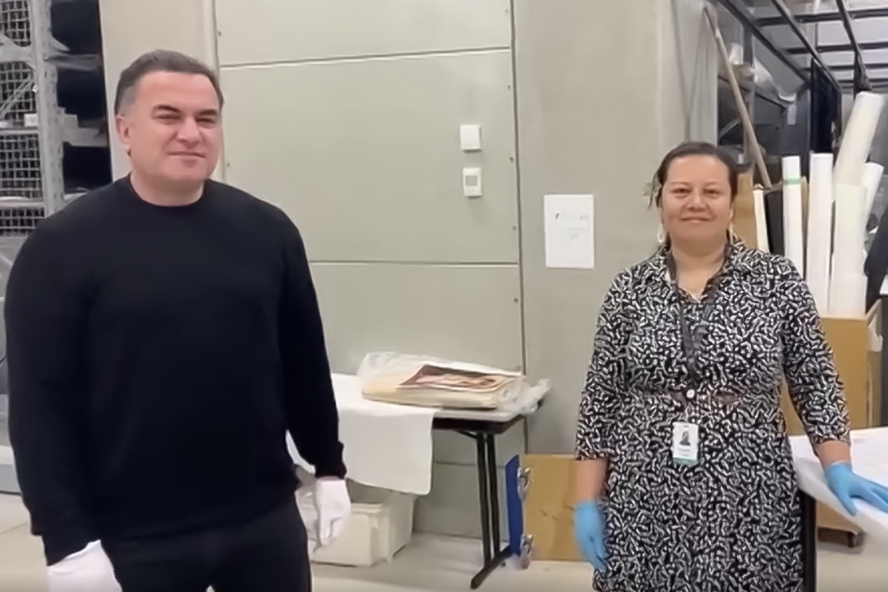 A man and a woman are standing in a museum store room. Both are looking at the camera.