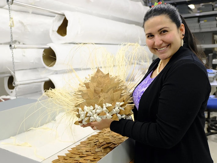 Kate Ngatokorua with the headpiece for Te Mana o te Ura, Pacific Culture Collections, Te Papa, 2022. Photo by Rachel Yates. Te Papa A woman is holding a headdress made of pandanus leaves and shells and smiling at the camera.