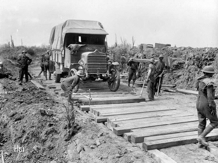 Members of the Pioneer Battalion laying a road in Messines, Belgium. Royal New Zealand Returned and Services' Association :New Zealand official negatives, World War 1914-1918. Ref: 1/2-012772-G. Alexander Turnbull Library, Wellington, New Zealand. /records/22563673 A black and white photo of men working in a war zone building a wooden road for a truck to pass.