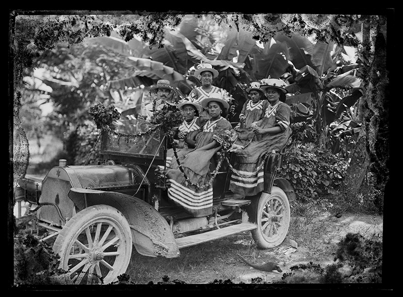 Five women and one man are dressed up and sitting in an open motor car, surrounded by trees.