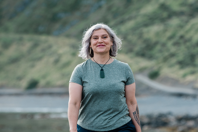 A woman with shoulder-length grey hair is stading outside in a t-shirt. She has a tattoo on her left arm and is wearing a piece of pounamu greenstone.