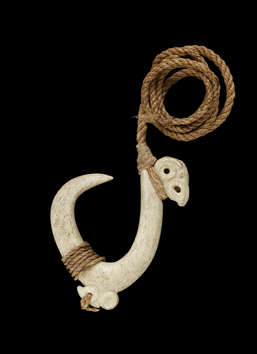 A bone fishhook carved with a pattern at one end and has a woven cord attached.