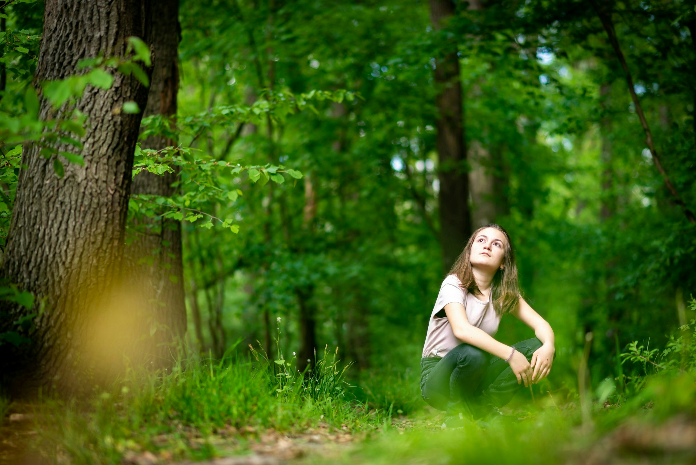 A young woman is sitting on the ground in a forest. She is looking up at the trees.