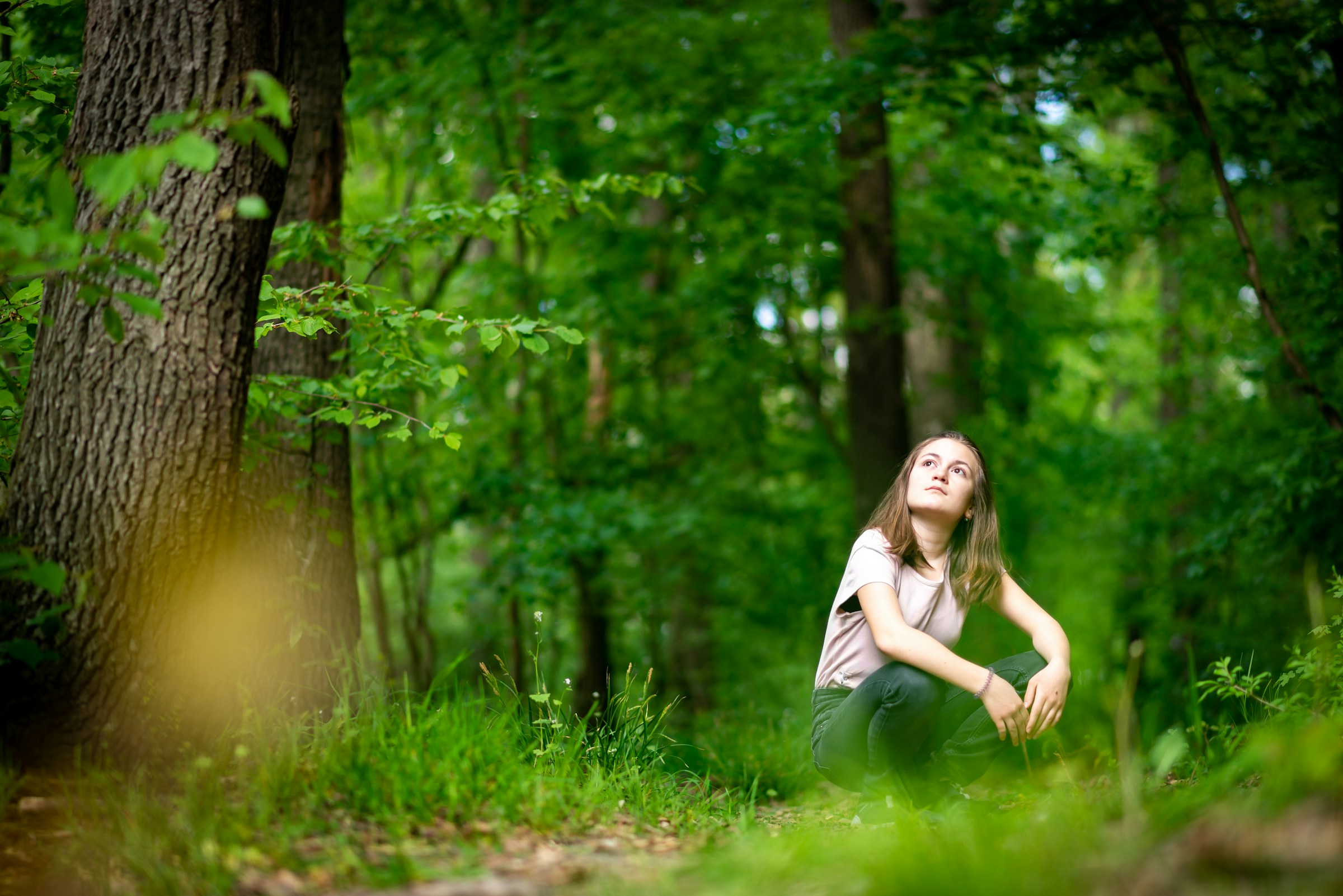 A young woman is sitting on the ground in a forest. She is looking up at the trees.