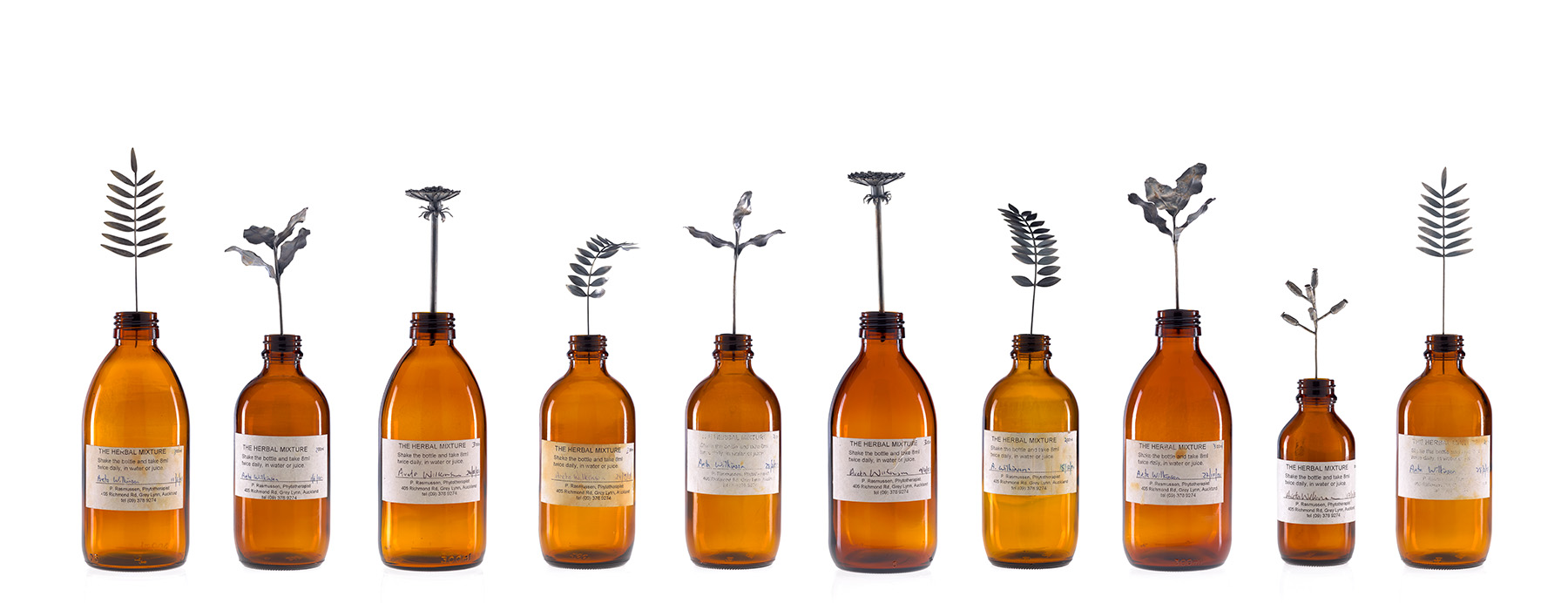 A photograph of ten brown bottles with labels lined up, Each bottle has a single silver leaf attached to a stem sticking out of it.