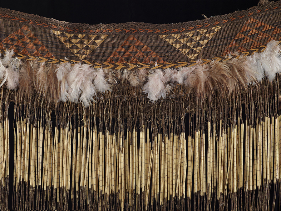 A piece of weaving that has a patterned band at the top, bird feathers in the next layer down and then long tassels.