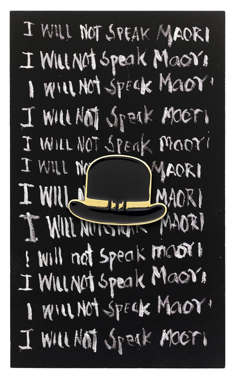 A black card with lines of white writing on it that says "I will not speak Maori". In the middle of the card is and enamel pin of a black bowler hat with a yellow band.