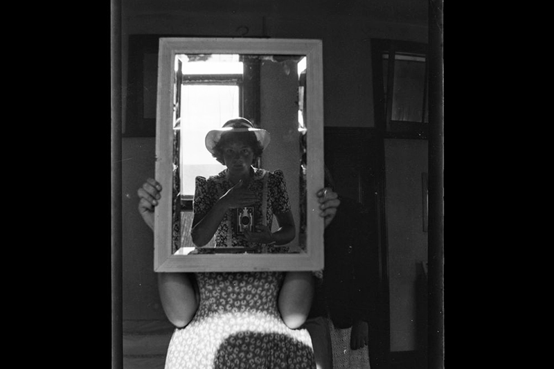 A black and white photograph of a woman sitting in a chair holding up a mirror which is reflecting another woman in a hat taking a photograph