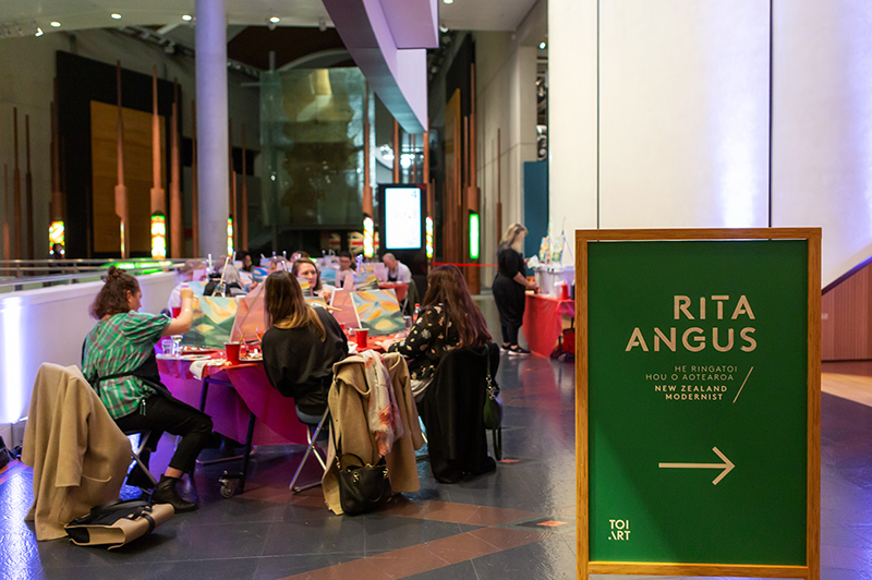 People sitting at tables in a large museum building. There is a sign in front of them directing people to the Rita Angus exhibition