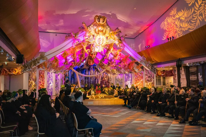 A lot of people are seated opposite each other in a large room. At the far end of the room is a large stage with ornate decoration and carving. It is lit up in pinks and oranges.