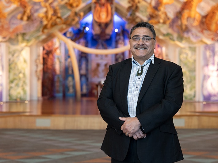 Kaihautū Arapata Hakiwai, 2021. Photo by Daniel Rose. Te Papa (174685) A man is standing in front of a colourful stage with his hands together in front of him.