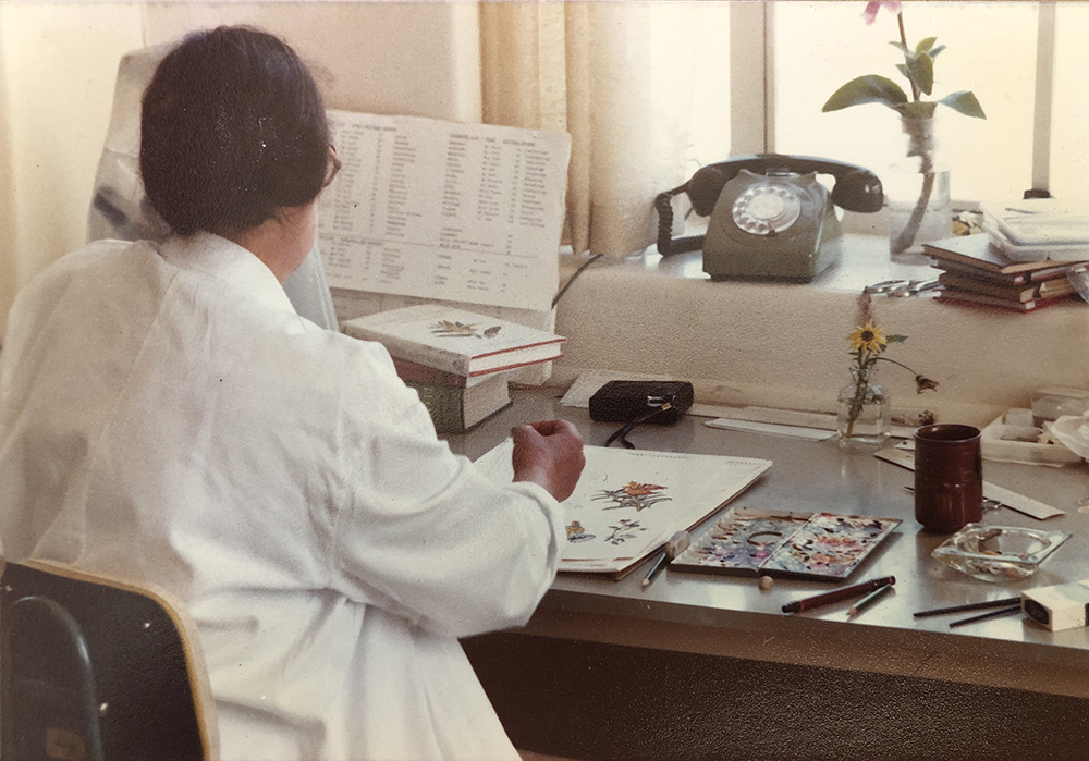 A woman sits at a desk and is painting flowers that are in a vase in front of her. She is in a white lab coat.