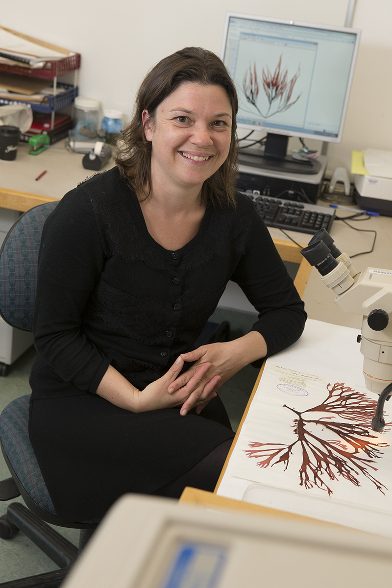 A woman in a black top is sitting at a desk. There is a page of algae in front of her. She is smiling at the camera.