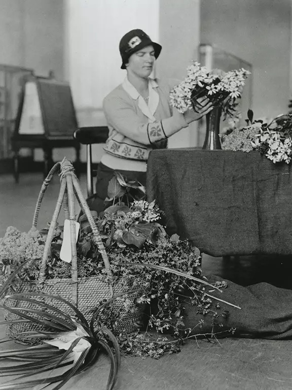 A black and white photo of a woman in a hat arranging flowers at a table.