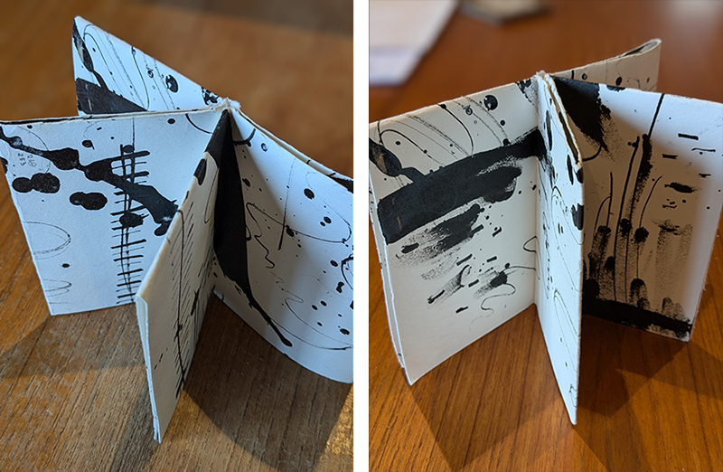 A small booklet with four pages of black text and blobs is open and standing up on a wooden table.