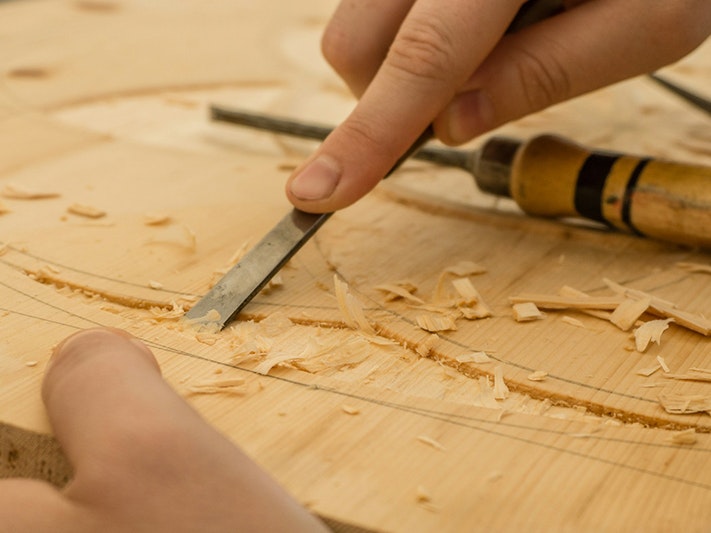 Photo by Dominik Scythe. Unsplash A closeup photo of two hands. One is holding a carving instrument ans is carving a piece of wood.