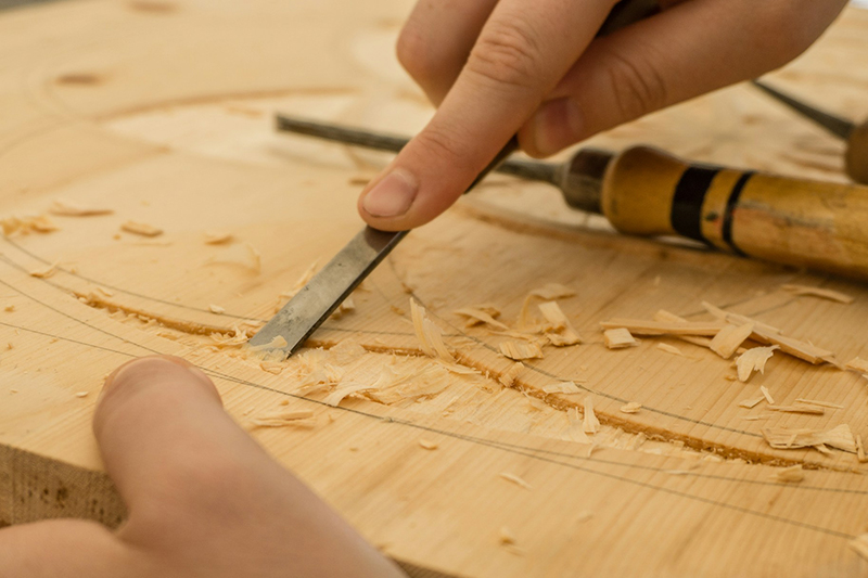 A closeup photo of two hands. One is holding a carving instrument ans is carving a piece of wood.