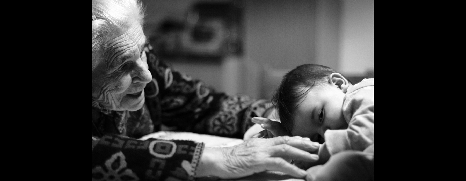 An elderly woman is smiling at a small baby child who is sleeping on its stomach