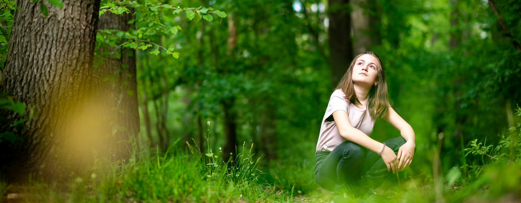 A young woman is sitting on the ground in a forest. She is looking up at the trees.