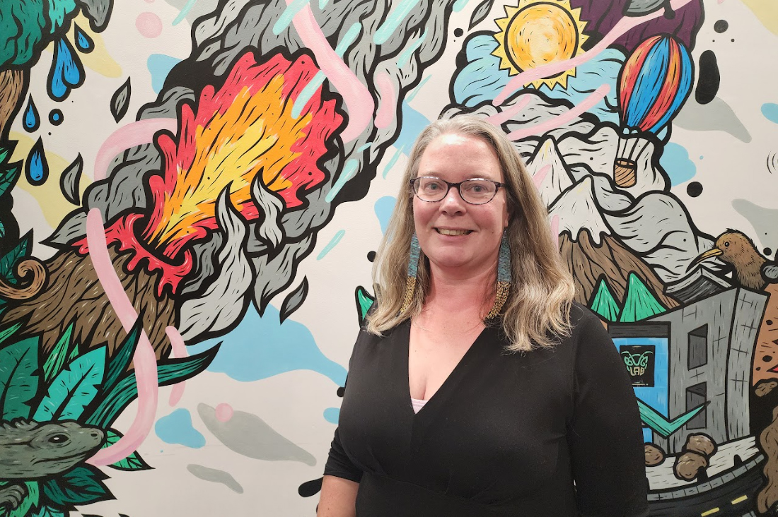 The top half of a woman with long hair standing in front of a colourful mural. She is smiling at the camera.