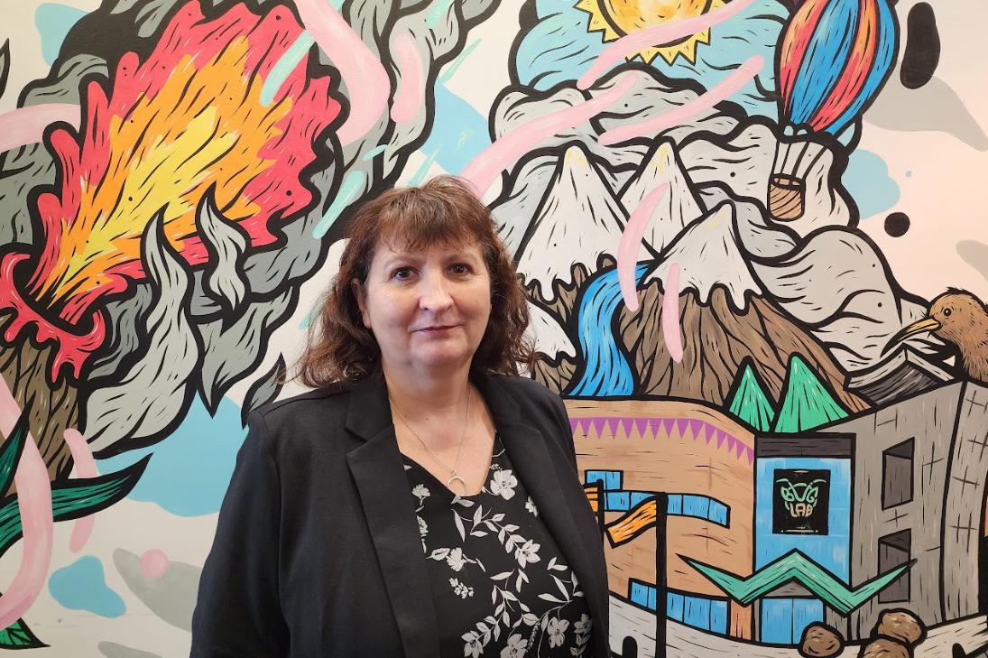 The top half of a woman with shoulder length hair standing in front of a colourful mural. She is smiling at the camera.