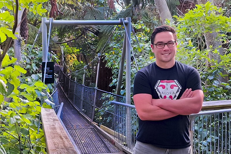 A man is standing on a walking bridge in the bush. He has is arms folded but is smiling at the camera.