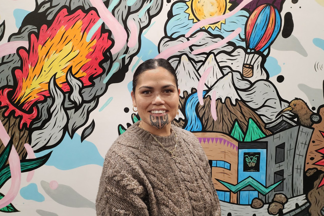 Head and shoulders of a woman with a chin tattoo and dark hair pulled back. She is standing in front of a colourful mural.