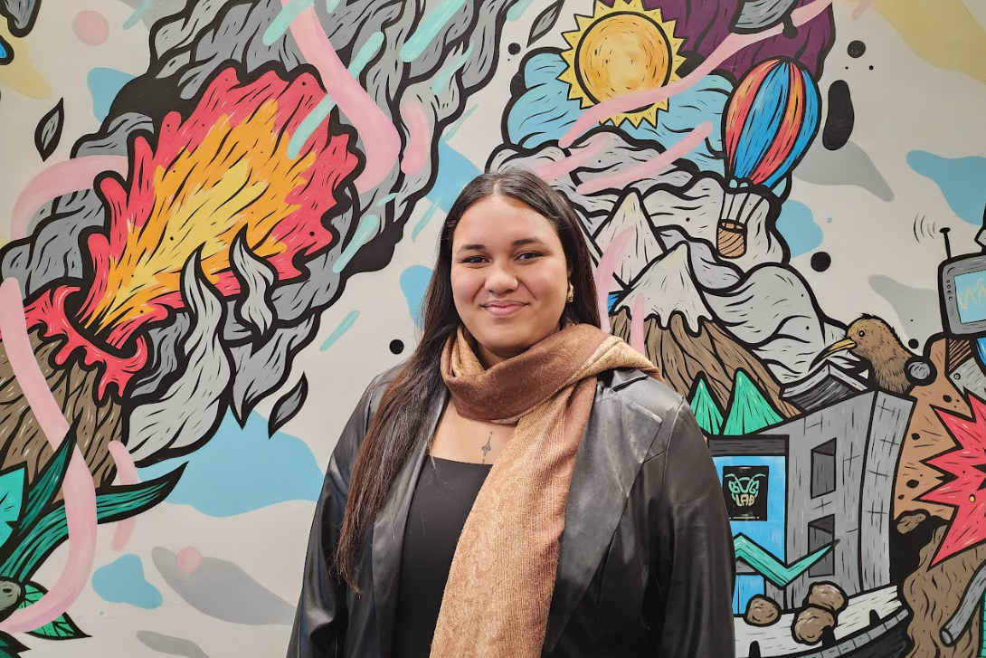 Top half of a woman with long hair, a long brown scarf and a leather jacket on. She is standing in front of a colourful mural.